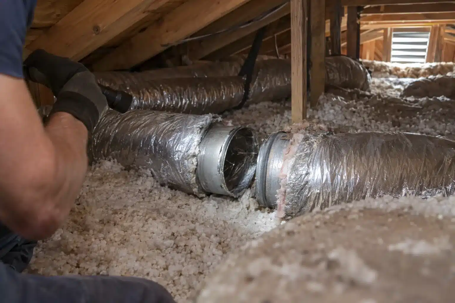 Gloved technician's hands holding a flashlight to inspect a loose flex duct joint in a hot Orlando home attic, with insulation and ductwork visible in the background