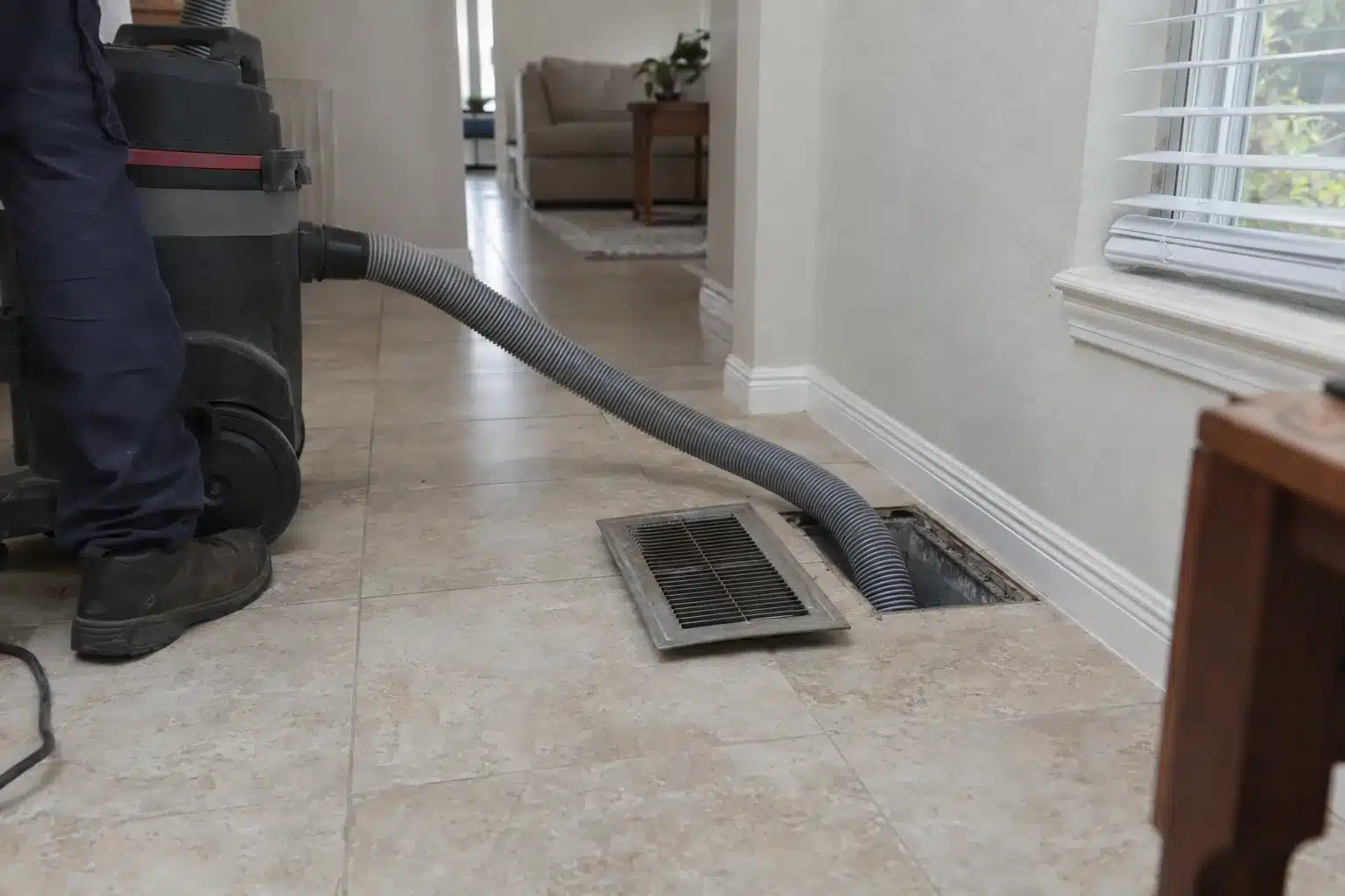 A portable duct cleaning vacuum hose inserted into an open floor register in a Florida home interior, with the register grille set aside on tile flooring nearby